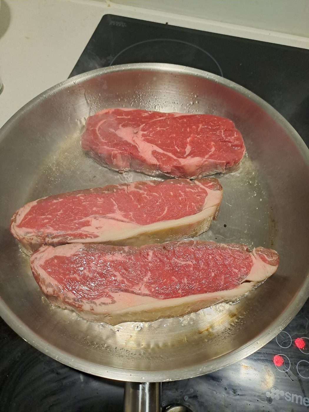 Three grass-fed steaks frying in ghee in a stainless steel pan