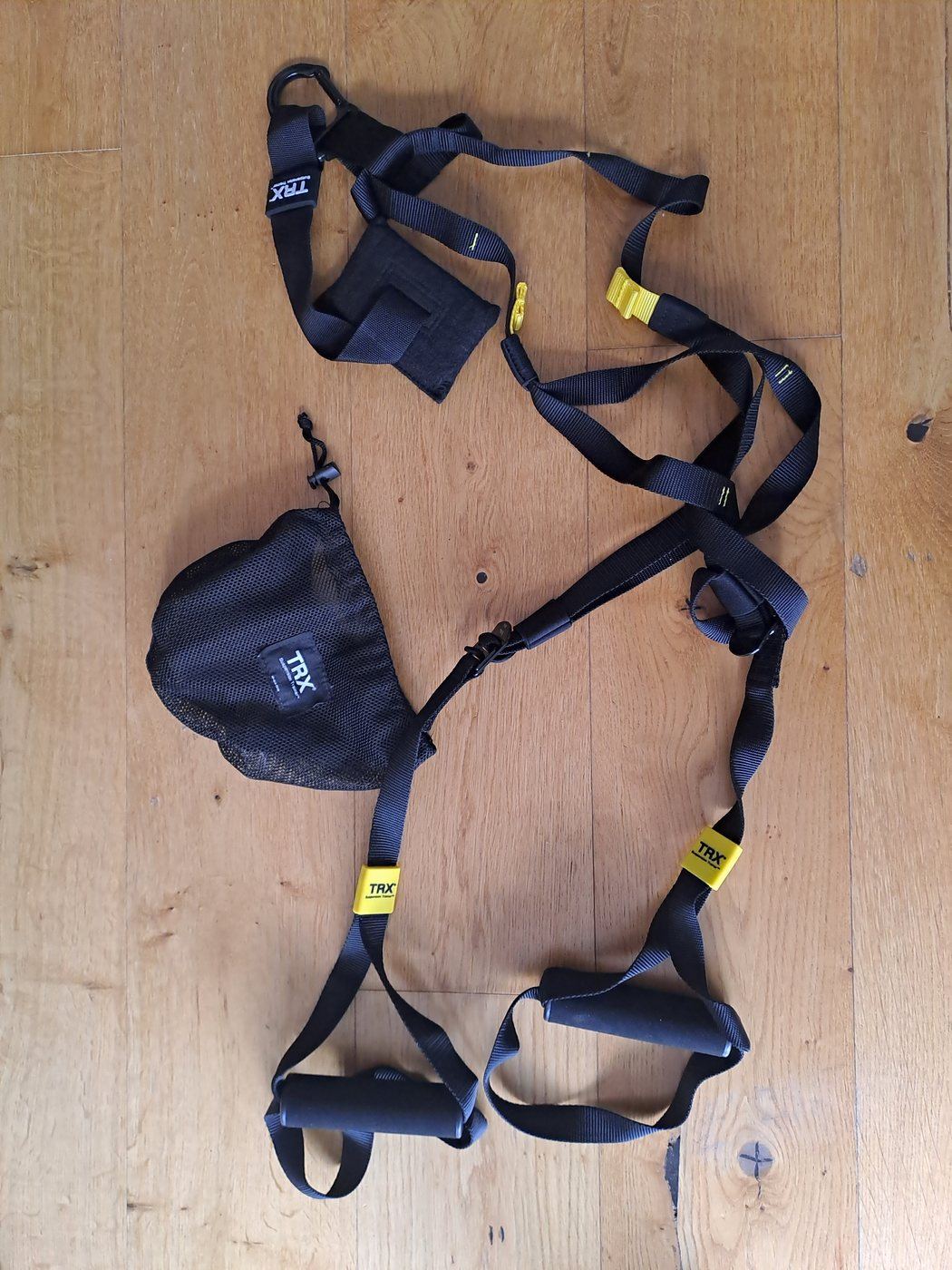 A TRX suspension trainer laid out on a wooden floor next to its mesh carry bag